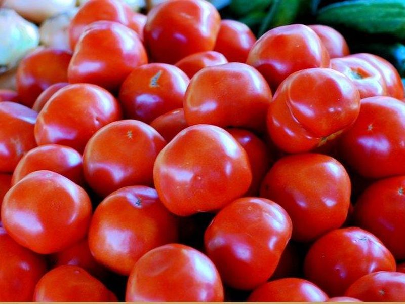 Ripe tomatoes piled at a market stand, with onions and cucumbers behind them