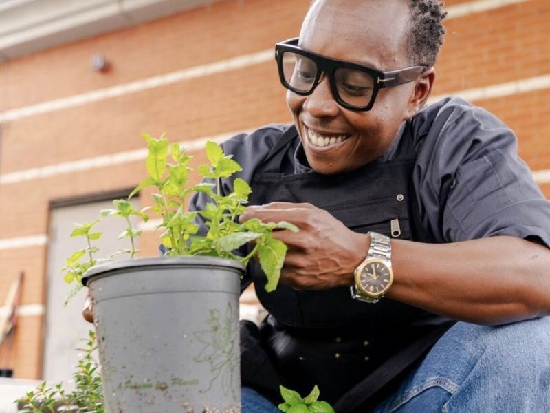 A chef crouching among potted herbs, holding a pot of basil